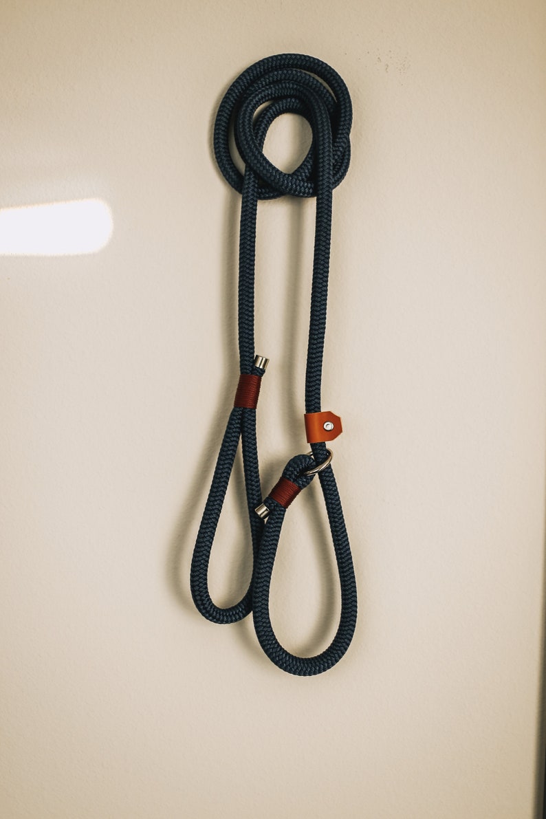 May include: A navy blue dog leash with a loop handle and a metal clasp. The leash is made of thick, braided rope and features brown leather accents. The leash is hanging on a white wall.