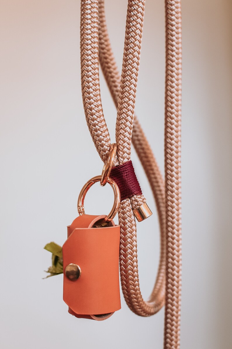 May include: A tan rope dog leash with a matching poop bag holder. The holder is made of orange leather and has a gold snap closure. The leash has a gold clasp and a burgundy accent.