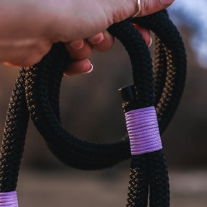 May include: Black rope dog leash with purple accents. The leash is made of thick, braided rope and features purple wrapping at the handle and near the clasp. The leash is held by a hand.