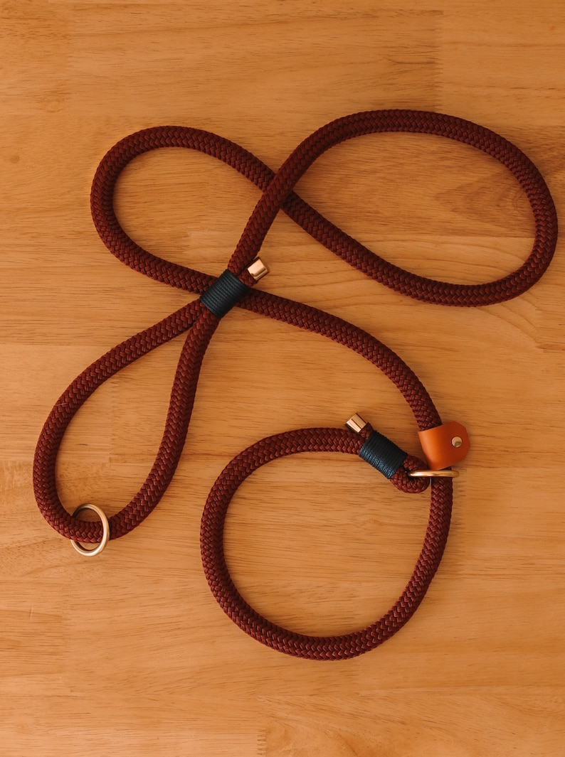 May include: A burgundy rope dog leash with a loop design. The leash features gold-colored metal accents, a dark teal accent, and a small tan leather detail. The leash is designed for easy handling and control.