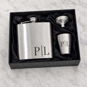 May include: A silver-toned stainless steel flask set presented in a black box with black fabric lining. The set includes a flask, a small funnel, and a shot glass. Both the flask and the shot glass are engraved with the letters P and L separated by a line.