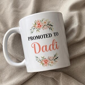 May include: A white ceramic mug featuring a floral design with pink and peach flowers above and below the black text PROMOTED TO and the coral-coloured text Dadi, which includes a small heart icon above the letter i on the front of the mug.