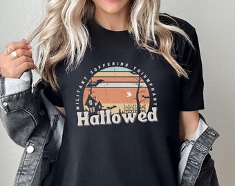 May include: Black t-shirt with a Halloween-themed graphic. The design features a haunted house, bats, and a graveyard under a sunset. The text reads "Hallowed" with the words "Militant Suffering Triumphant" arched above.