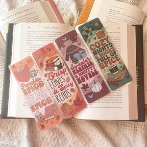 Könnte beinhalten: Vier farbenfrohe Lesezeichen mit Herbstmotiven. Jedes Lesezeichen zeigt ein anderes Design mit Herbstfarben, Blättern und Büchern. Der Text auf den Lesezeichen lautet "Pumpkin Spice and Books with Even More Spice", "Crisp Leaves & Spicy Reads", "Sweater Weather & Smutty Novels" und "Cozy Nights & Books Full of Spice".