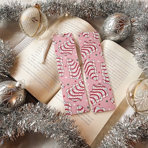 May include: Two pink and white Christmas tree patterned bookmarks with a tassel. The bookmarks feature a repeating pattern of Christmas trees with red and white stripes and green leaves. The background is pink with white stars.