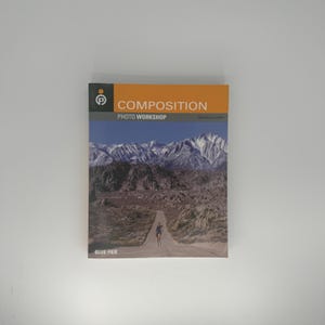 May include: A book titled "COMPOSITION PHOTO WORKSHOP" with a landscape photo on the cover. The photo shows a person walking down a road towards snow-capped mountains. The book has a white background and the text "BLUE FIER".