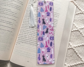 May include: A pink and purple bookmark with a black cat, cauldron, crystals, and other witchy symbols. The bookmark has a silver tassel.