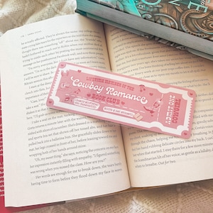 May include: A pink bookmark with the text "Lifetime Member of the Cowboy Romance Book Club" and "Admit One Cowgirl." The bookmark has a ticket design with cowboy-themed illustrations and is placed on an open book.