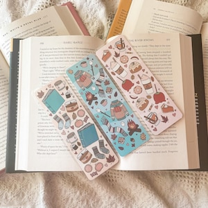 May include: Three colorful bookmarks with autumn-themed illustrations. The bookmarks feature a variety of fall-inspired designs, including a cozy sweater, a mug of hot cocoa, a pumpkin pie, and a book. The bookmarks are printed on a clear, plastic-like material.