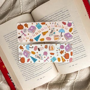 Peut inclure: Deux marque-pages rectangulaires avec un fond blanc et des illustrations colorées sont affichés ouverts sur un livre. Les marque-pages présentent des images de livres, de citrouilles, de tasses à café et le texte "La La La".