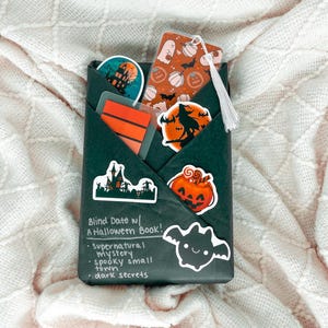 Puede incluir: Un paquete negro envuelto con pegatinas temáticas de Halloween y un marcapáginas. Las pegatinas presentan una casa embrujada, una calabaza, una bruja y un murciélago. El marcapáginas tiene un diseño de calabaza y fantasma. El texto en el paquete dice "Blind Date w/ A Halloween Book!"