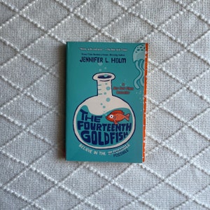 Könnte beinhalten: Ein blaugrüner Buchumschlag mit dem Titel "The Fourteenth Goldfish" in weißer Schrift. Der Buchumschlag zeigt einen Cartoon-Goldfisch in einem Becherglas mit dem Text "Believe in the Impossible Possible" am unteren Rand.