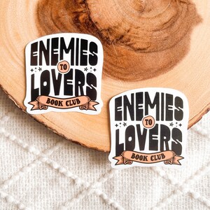 May include: Two black and white stickers with the text "Enemies to Lovers Book Club" on a light brown wooden surface. The stickers have a peach colored banner with the text "Book Club".