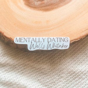 Op de afbeelding: Een witte sticker met de tekst "MENTALLY DATING Wells Whitaker" in een mix van sans-serif en script letters. De sticker ligt op een houten oppervlak met een lichtgekleurde, getextureerde achtergrond.