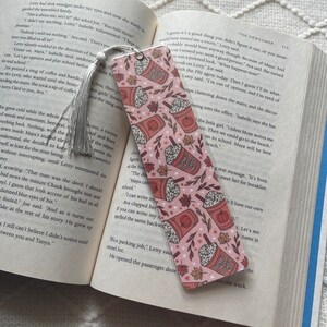 May include: A pink bookmark with a fall-themed design featuring multiple illustrations of pumpkin spice lattes with whipped cream and sprinkles. The bookmark also has small illustrations of fall leaves and stars.