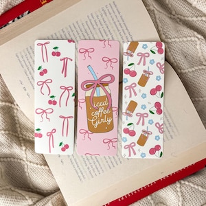 May include: Three decorative bookmarks with cherry and bow patterns. One bookmark features an iced coffee illustration with the text "Iced Coffee Girly". The bookmarks are on a book.