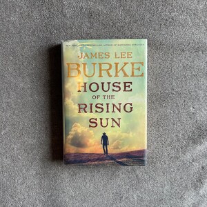 Puede incluir: Una portada de libro con un fondo de puesta de sol y una silueta de una persona caminando por un camino de tierra. El título del libro es "House of the Rising Sun" de James Lee Burke.