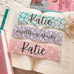 May include: Three clear bookmarks with a floral design. The top bookmark has a white background with green leaves and white daisies. The middle bookmark has a purple background with white daisies. The bottom bookmark has a pink background with white daisies. All three bookmarks have the name "Katie" written in black cursive. The middle bookmark also has the text "@spellboundbooks" written in black cursive.