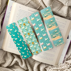 May include: Four decorative bookmarks with swan and summer-themed designs. One bookmark features swans and ice cream popsicles, another with sunflowers, and two with swans on inner tubes. The bookmarks are on an open book.