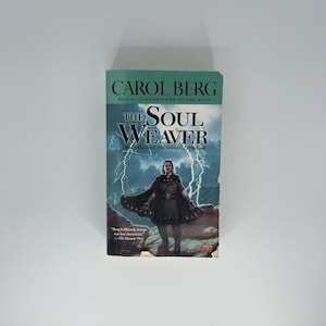 May include: A paperback book titled "The Soul Weaver" by Carol Berg. The cover features a woman in a dark cloak against a backdrop of lightning and mountains. The book is part of "The Bridge of D'Aruath" series.