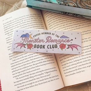 May include: A bookmark with the text "PROUD MEMBER OF THE Monster Romance BOOK CLUB" in pink and black. The design includes a crescent moon, clouds, stars, roses, and bat wings.