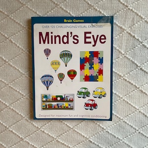 Op de afbeelding: Een boek getiteld "Mind's Eye" met illustraties van luchtballonnen, legpuzzels en auto's. De omslag bevat de tekst "Brain Games" en "Over 125 Challenging Visual Exercises". Het boek is ontworpen voor plezier en cognitieve conditionering.