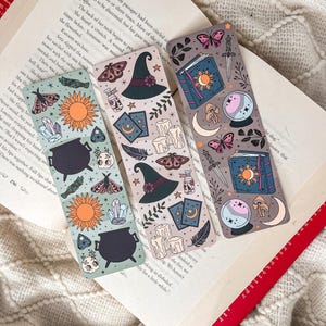May include: Three decorative bookmarks with mystical illustrations. One features a cauldron, sun, and crystals. Another shows a witch's hat, candles, and cards. The third has a book, crystal ball, and butterflies. Each bookmark is approximately 7 inches long.