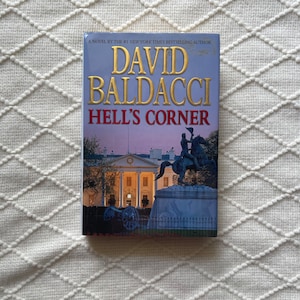 May include: A paperback book cover with a white background and a blue sky. The title "Hell's Corner" is in large, bold, red letters. The author's name, "David Baldacci", is in large, bold, black letters. The cover features a statue of a man on horseback in front of a white building.