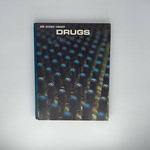 May include: A hardcover book titled "DRUGS" from the "LIFE SCIENCE LIBRARY." The cover features a dark, textured background with a pattern of blue and black cylindrical shapes. The book has a blue spine.