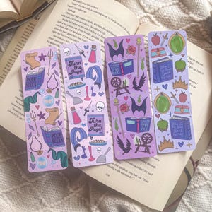May include: Four purple bookmarks with whimsical illustrations. Each bookmark features unique designs, including books, potions, and text that reads "Turn the Page!" and "Poor Souls." The bookmarks are on top of an open book.