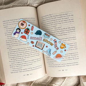 Puede incluir: Un marcap&aacute;ginas azul claro con el texto "I smell snow" e ilustraciones de invierno. El marcap&aacute;ginas presenta im&aacute;genes de un mu&ntilde;eco de nieve, un gorro y patines de hielo. Est&aacute; sobre un libro abierto.