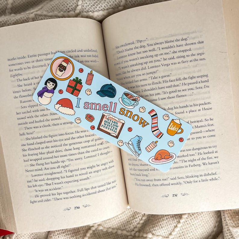 Puede incluir: Un marcap&aacute;ginas azul claro con el texto "I smell snow" e ilustraciones de invierno. El marcap&aacute;ginas presenta im&aacute;genes de un mu&ntilde;eco de nieve, un gorro y patines de hielo. Est&aacute; sobre un libro abierto.