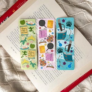 May include: Three decorative bookmarks with illustrated designs. One green bookmark features a frog, eggs, and the text "There's magic in this Book." Another pink bookmark has a frying pan, paint palette, and the text "Best Book Ever." The third blue bookmark shows a bow and arrow, bear paw prints, and a book.