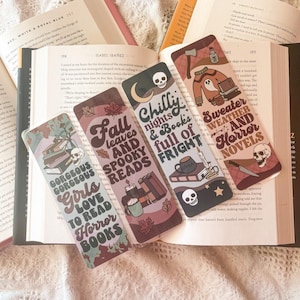 May include: Four colorful bookmarks with autumnal themes. Each bookmark features a different design with illustrations of books, skulls, and other spooky elements. The text on the bookmarks reads "Fall leaves and spooky reads", "Chilly nights & books full of fright", "Gorgeous Gorgeous Girls to love to read horror books", and "Sweater weather and horror novels".