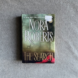 以下が含まれることがあります： ノラ・ロバーツ著「The Search」というタイトルのペーパーバック。表紙には犬のシルエットと著者の名前が白で描かれた森の風景が描かれています。本は灰色の背景に置かれています。