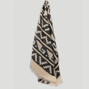 May include: A black and white patterned throw blanket with fringe on the edges. The blanket is folded and hanging on a white wall.