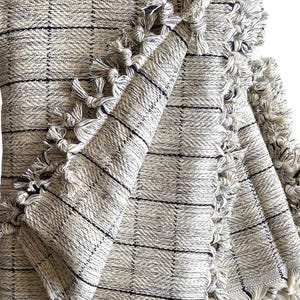 May include: A cream-colored woven throw blanket with black stripes and fringe trim. The blanket is folded in half and the fringe is visible on both sides.