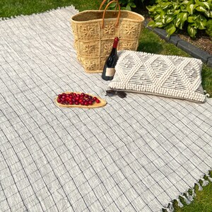 May include: A white and gray checkered picnic blanket with fringe edges. A woven basket, a bottle of wine, a plate of cherries, and a pillow are on the blanket.