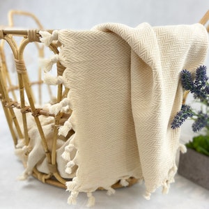 May include: A cream-colored woven throw blanket with fringe trim is draped over a wicker basket. The blanket has a herringbone pattern.