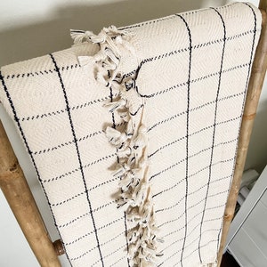 Könnte beinhalten: Eine weiße Decke mit schwarzen Streifen und Fransen. Die Decke ist gefaltet und über einem Bambusgestell drapiert.