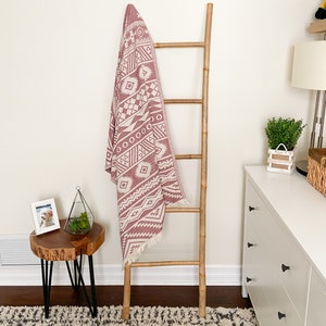 Peut inclure: Une couverture en coton rose et blanc à motifs drapé sur une échelle en bois. L'échelle est posée sur un tapis blanc à motifs noirs et blancs.