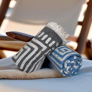 Pu&ograve; includere: Due asciugamani da spiaggia, uno grigio con motivi geometrici bianchi e uno blu con motivi geometrici bianchi, sono arrotolati e posizionati su una superficie marrone chiaro.