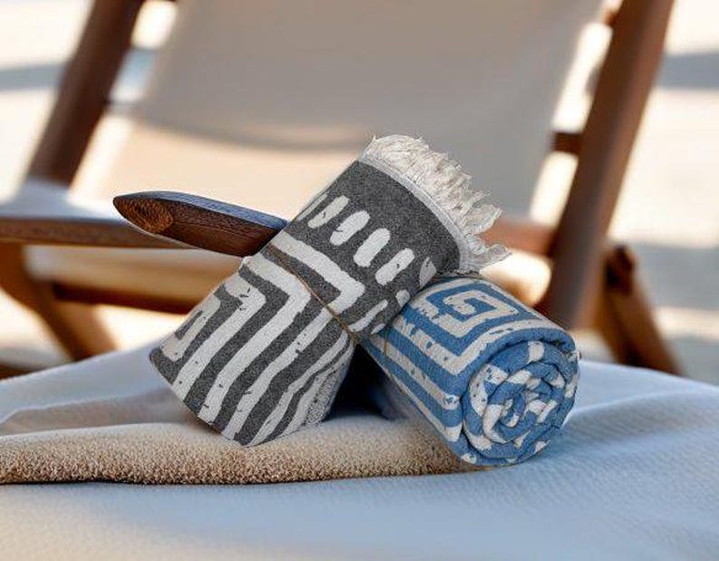 Pu&ograve; includere: Due asciugamani da spiaggia, uno grigio con motivi geometrici bianchi e uno blu con motivi geometrici bianchi, sono arrotolati e posizionati su una superficie marrone chiaro.