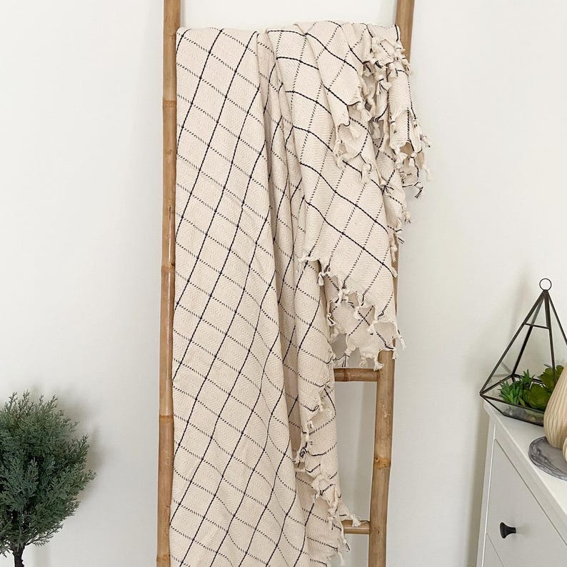 May include: A cream-colored throw blanket with a black diamond pattern and fringe edges. The blanket is draped over a wooden ladder.
