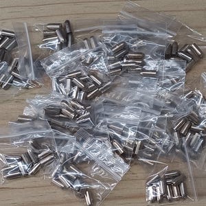 May include: Multiple small clear plastic bags filled with silver-colored metal objects. The objects appear to be cylindrical with rounded ends. The bags are scattered across a surface, suggesting a bulk quantity of the items.