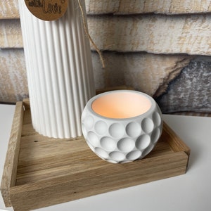 Könnte beinhalten: Weiße Keramikvase mit einem braunen Etikett mit der Aufschrift "Handmade with Love". Die Vase steht auf einem Holztablett mit einem weißen Kerzenhalter aus Keramik mit strukturierter Oberfläche.
