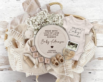Könnte beinhalten: Ein rundes Schild aus weißem Holz mit dem Text "Made with lots of love and a little science Baby Adamson Coming April 2024". Das Schild ist umgeben von einem Baby-Strampler, einem Paar Stiefelchen, einem Holzblock, der "BABY" buchstabiert, einem kleinen Stofftier und einem weißen Blumenarrangement.