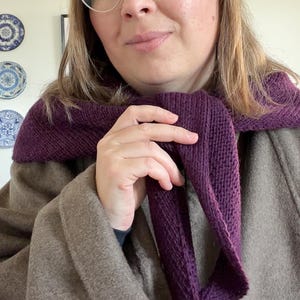 May include: A deep purple knitted scarf with a textured stitch pattern draped around the neck, worn over a solid brown coat. The scarf features a consistent knit construction and is tied loosely in the front, showcasing the dark plum color.