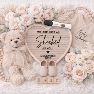 Puede incluir: Anuncio de bebé en tonos neutros con un oso de peluche, flores y un letrero en forma de corazón con el texto "WE ARE JUST AS SHOCKED AS YOU!" y "DECEMBER 2026". Incluye zapatos de bebé, un body y una prueba de embarazo.
