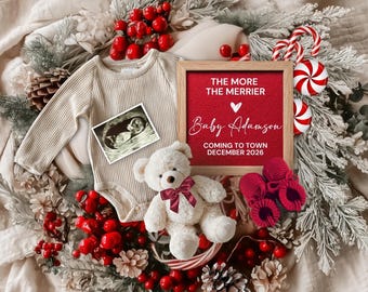 May include: A festive pregnancy announcement with a cream-colored onesie, ultrasound image, and a teddy bear with a red bow. A framed sign reads "The More The Merrier Baby Adamson Coming to Town December 2026". Red booties and candy canes complete the holiday theme.